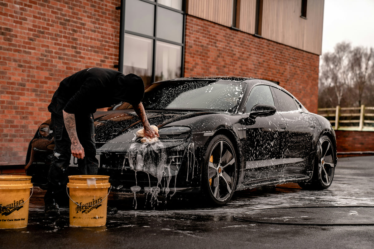 En person som vasker en svart Porsche Taycan med en svamp og såpeskum, med to Meguiars-bøtter i nærheten.