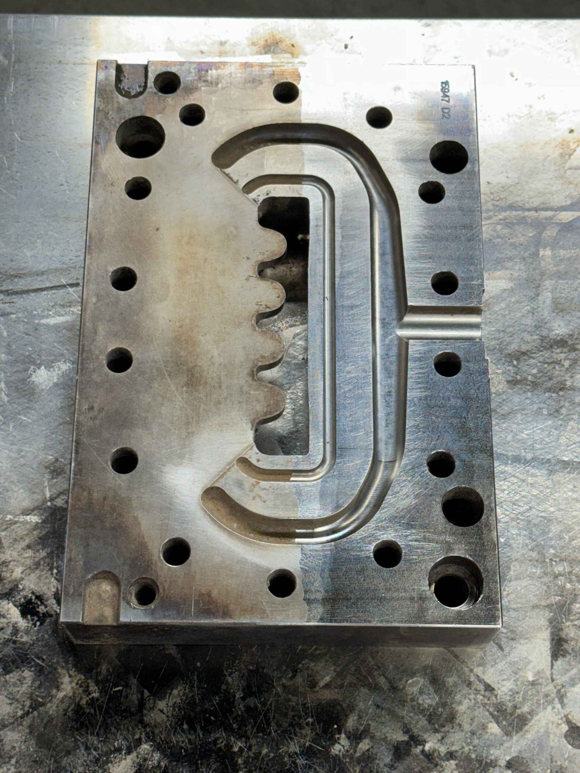 En rektangulær metallform med flere hull og en utskjæring i midten ligger på en riflet metalloverflate.