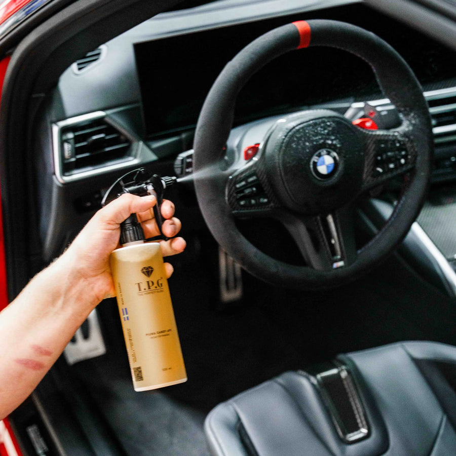 En hånd holder en TPG Piuma Candy APC-flaske fra The Perfect Gloss i en BMW med svart skinnsete og ratt.